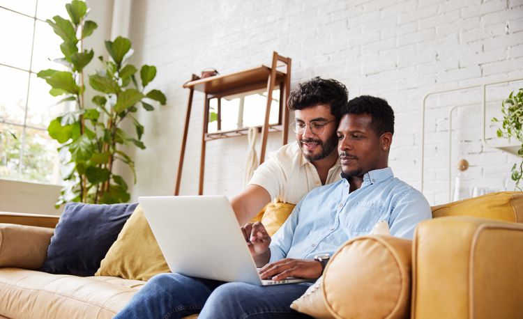 A man sits on a couch with a laptop while another man stands behind him looking over his shoulder and pointing at the laptop